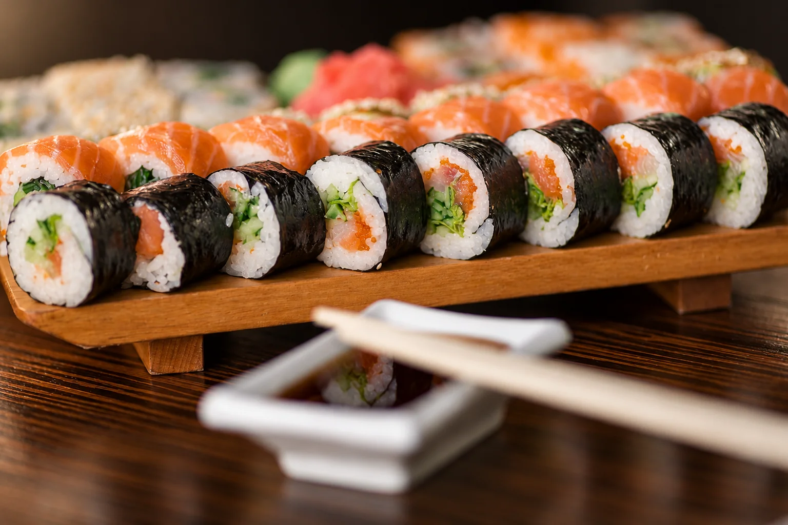 Une rangée de makis garnis de saumon et de verdure sur une planche en bois, avec une coupelle blanche de sauce soja et des baguettes au premier plan.